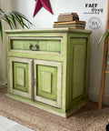Dixie Belle Paint Limeade Chalk Mineral Paint is the perfect paint for any DIY project!
A Cabinet painted with a mix of Limeade and Evergreen Chalk Mineral Paint, there are off white areas around the edges of the door and the drawer. Staged with a stack of books on top, as well as a ladder with multiple colors hanging on it to the right side of the piece.