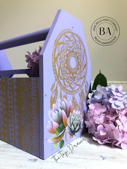 Dixie Belle Paint Lucky Lavender Chalk Mineral Paint is the perfect paint for any DIY project!
Wooden toolbox painted in Lucky Lavender Chalk Mineral Paint with the Mudcloth Stencil on the front of the box and Western Boho - Silkscreen Stencil on the side. Staged with pink & purple flowers.