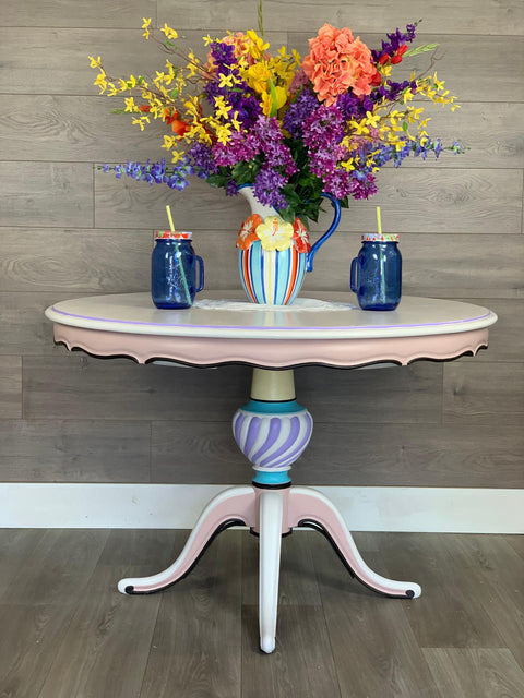 Dixie Belle Paint Lucky Lavender Chalk Mineral Paint is the perfect paint for any DIY project!
Table painted in white and pink with Lucky Lavender Chalk Mineral Paint  used as an accent color along the base.. Staged with a blue,yellow,and orange pitcher with multicolored flowers and two blue mason jar cups  on each side.