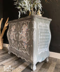 Dixie Belle Paint Mason Dixon Gray Chalk Mineral Paint is the perfect paint for any DIY project! Chalk Painted gray Side table with Stencil squares on the side and florals on the front of drawers, with white painted accents all around it. Staged with some plants on top.