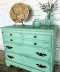 Dixie Belle Paint Mint Julep Chalk Mineral Paint is the perfect paint for any DIY project!
Mint distressed chalk painted dresser with gold hardware. Staged with two glass vases with plants inside and a wicker basket in a plate holder.