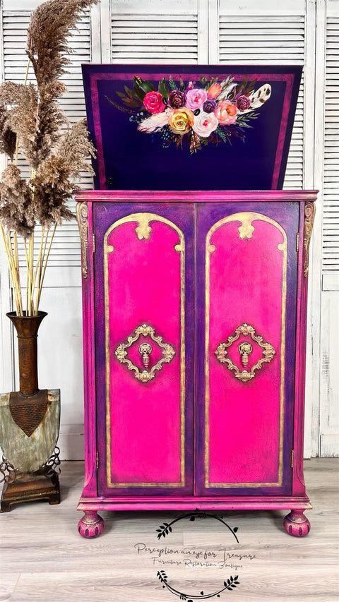 Dixie Belle Paint Peony Chalk Mineral Paint is the perfect paint for any DIY project! Pink chalk painted open top cabinet blended with purple around the doors in front with gold on the trim and a floral rub on transfer on the top of the cabinet door. Staged next to faux wheat plants in a vase.