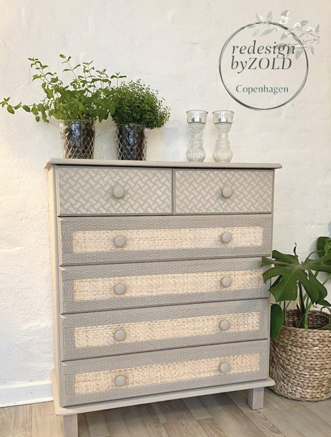 Dixie Belle Paint Putty Chalk Mineral Paint is the perfect paint for any DIY project! A muted taupe colored dresser featuring 2 short drawers next to each other on top with a basket weave stencil and 4 long skinny drawers underneath with a rattan like pattern surrounded by various potted greenery.