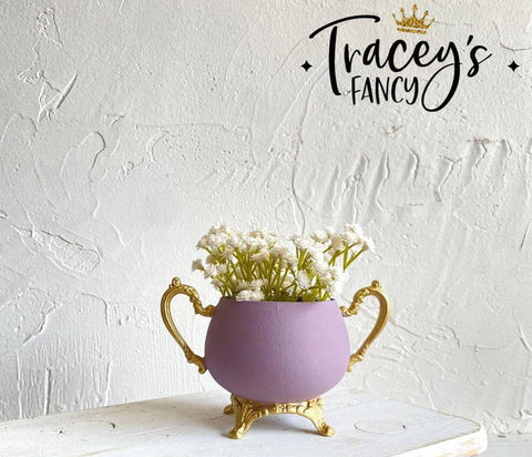 Purple chalk painted pitcher with gold legs and handles. Staged with white flowers inside, on a white painted distressed stool.