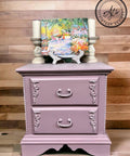 Purple chalk painted nightstand with scrolls on the sides of the drawers. Staged with two large candlesticks and a colorful painting on top.