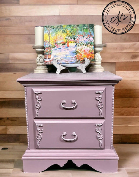 Purple chalk painted nightstand with scrolls on the sides of the drawers. Staged with two large candlesticks and a colorful painting on top.