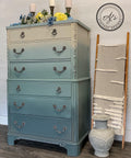 Dixie Belle Paint Stormy Seas Chalk Mineral Paint is the perfect paint for any DIY project!
Tall dresser Painted tan ombre to Stormy Seas with silver hardware. Staged with blue,yellow,white flowers along the top of dresser and three bronze candle holders in the back.