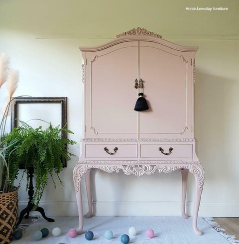 Dixie Belle Paint Tea Rose Chalk Mineral Paint is the perfect paint for any DIY project!
A large drink cabinet painted in Tea Rose Chalk Mineral Paint with dark hardware and a black tassle on the door handle. Staged on top of a white fringe rug, party lights on the floor and greenery.