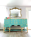Dixie Belle Paint The Gulf Chalk Mineral Paint is the perfect paint for any DIY project!  A blue chalk painted dresser with gold legs, knobs and decorative scrolls sitting on the floor in front of a wall with a large gold mirror.