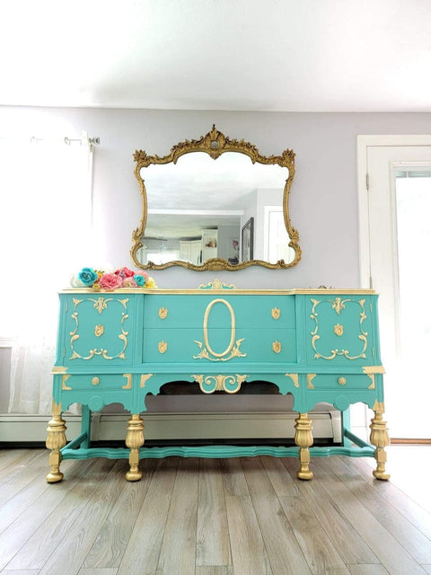 Dixie Belle Paint The Gulf Chalk Mineral Paint is the perfect paint for any DIY project!  A blue chalk painted dresser with gold legs, knobs and decorative scrolls sitting on the floor in front of a wall with a large gold mirror.