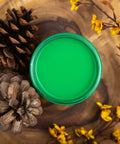 Dixie Belle Paint Tree Frog Green Chalk Mineral Paint is the perfect paint for any DIY project!  An open jar of bright green Dixie Belle Tree Frog Green Chalk Mineral Paint sitting on a piece of wood with pine cones and branches with flowers.