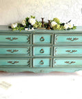 Dixie Belle Paint Vintage Duck Egg Chalk Mineral Paint is the perfect paint for any DIY project!  A greenish blue chalk painted dresser with gold handles sits on a white floor in front of a white wall.  White flowers with greenery and gold candle sticks sit on top of the dresser.  An open book with loose pages sits on the floor to the left of the dresser.
