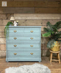 Dixie Belle Paint Vintage Duck Egg Chalk Mineral Paint is the perfect paint for any DIY project!  A bluish green chalk painted dresser sitting on a brown wooden floor, in front of a wall with brown wooden paneling.  A small lamp, white angel figuring, white candle on a brown candlestick and a green plant sit on top of the dresser.  A green plant inside a brown basket sit on top of a small wooden stool to the right of the dresser, and a gray furry rug lies on the floor.