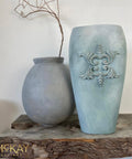 Dixie Belle Paint Vintage Duck Egg Chalk Mineral Paint is the perfect paint for any DIY project!  A small rounded gray chalk painted clay pot and a grayish-blue blended chalk painted clay pot with ornamental decorations sit on a piece of wood, in front of a white wall.  A stick is coming out of the small gray pot.