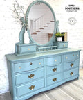 Dixie Belle Paint Vintage Duck Egg Chalk Mineral Paint.  A blue chalk painted dresser with a large oval mirror on top of it and bronze & white handles.  A white vase holding brown sticks with white flowers and greenery sits to the left of the mirror on the dresser.  A white pot holding a green plant and 3 black books sit to the right of the mirror.  The dresser sits on a gray and white striped rug, in front of a white brick wall.