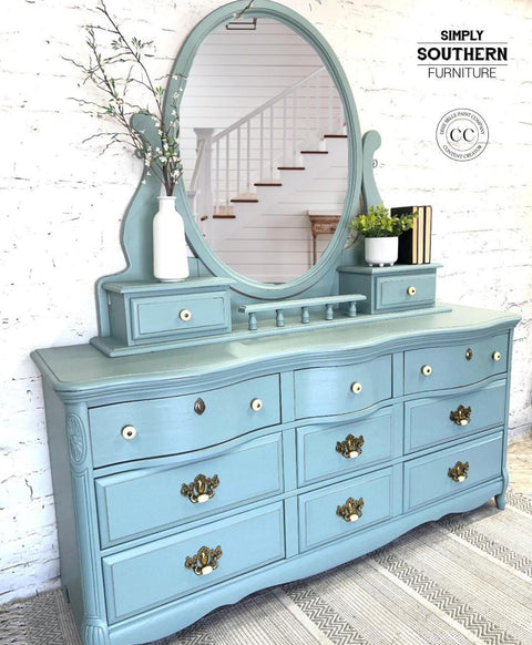 Dixie Belle Paint Vintage Duck Egg Chalk Mineral Paint.  A blue chalk painted dresser with a large oval mirror on top of it and bronze & white handles.  A white vase holding brown sticks with white flowers and greenery sits to the left of the mirror on the dresser.  A white pot holding a green plant and 3 black books sit to the right of the mirror.  The dresser sits on a gray and white striped rug, in front of a white brick wall.