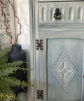 Dixie Belle Paint Whitewash Glaze. A gray chalk painted cabinet with bronze hardware has Whitewash Glaze applied to the front of it.  To the left of the cabinet is a black chalk painted vase holding two brown sticks and a green plant.