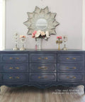 Dixie Belle Paint Yankee Blue Chalk Mineral Paint is the perfect paint for any DIY project!  A Yankee Blue chalk painted dresser with gold hardware sits on a light brown wooden floor, in front of a gray wall with white trim.  A round mirror with silver flower petals hangs on the wall above the dresser.  Several silver candlesticks with clear hanging ornaments sit on top of the dresser, along with pink and white roses inside of a clear glass vase.