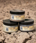 Three jars of Dixie Belle Dixie Dirt in different shades are stacked on dirt, with black lids and tan labels, each containing 118 ml (4 oz) of product. The background is a textured soil surface.