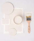 White paint samples arranged artfully with a jar of paint and a wooden-handled paintbrush on a white background. The samples include circular and rectangular pieces of paper.
