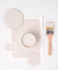 A round paint can filled with beige paint is surrounded by paint swatches and circles on a white background. A wooden-handled paintbrush with paint on its bristles is placed on the right side of the arrangement.