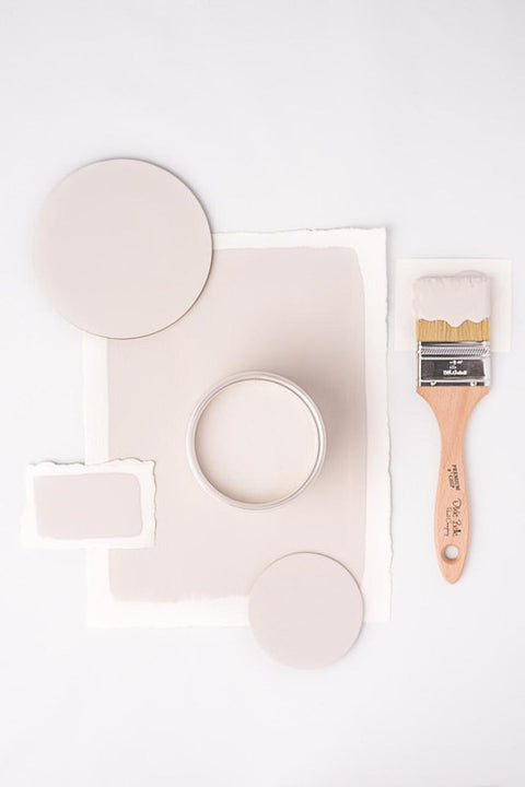 A round paint can filled with beige paint is surrounded by paint swatches and circles on a white background. A wooden-handled paintbrush with paint on its bristles is placed on the right side of the arrangement.