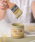 Person holding a brush with greenish paint dripping over an open jar labeled "Dixie Belle Chalk Mineral Paint Olive Wood," placed on a surface with dried flowers nearby.