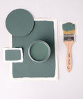 A flat lay image featuring a jar of green paint, a paintbrush with a wooden handle partially covered in green paint, and green paint swatches on white paper, all against a light background.