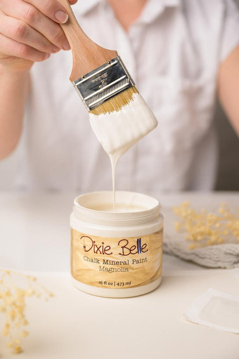A person dips a paintbrush into a jar of Dixie Belle chalk mineral paint in Magnolia. The paint appears creamy and white, dripping from the brush. The jar is on a white surface with decorative elements nearby.