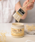 A person holds a brush coated with pale cream-colored paint above an open jar labeled "Dixie Belle Chalk Mineral Paint Ecru." The jar is on a white surface with dried flowers nearby. The background is softly blurred, focusing on the paint and brush.