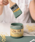 A person holds a paintbrush dripping with green paint above an open jar labeled "Dixie Belle Chalk Mineral Paint Blue Grass." The paintbrush is partially covered in matching green paint. Dry flowers and a fabric piece are nearby.