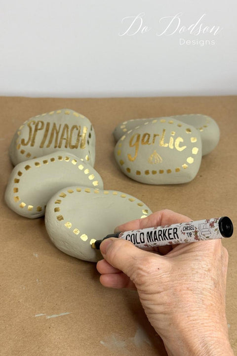 Hand writing on rocks with a gold marker, labeled 'Spinach' and 'Garlic'.