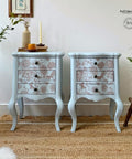 A pair of light blue vintage nightstands with three drawers each, adorned with floral patterns in light brownish red. The stands are placed on a woven rug, with potted plants, a candle, and books on top. A framed picture hangs on the wall above the left stand.