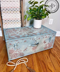 A decorated blue trunk with floral and dragonfly patterns holds a potted plant. A long, beaded wooden garland is arranged in front on the wooden floor. A rustic striped towel hangs from a ladder in the background, and a watermark logo reads "AJ's Vintage Designs".