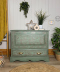 A stylish, vintage green dresser with floral patterns and brass handles is surrounded by various decorative items. To the left, a ladder with yellow blankets and hanging baubles leans against the wall. Several plants and a circular woven rug complete the cozy scene.