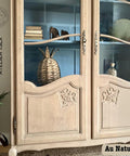 A rustic wooden cabinet with glass doors showcases decorative items, including stacked books, a woven basket, and small ornaments. The cabinet features ornate designs and is placed against a blue-painted interior background. Text reads "Au Naturel.