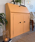 An orange-colored wooden write-on cabinet with a slanted top and two doors, styled in a home interior next to a chair, plants, and a woven basket candle holder.
