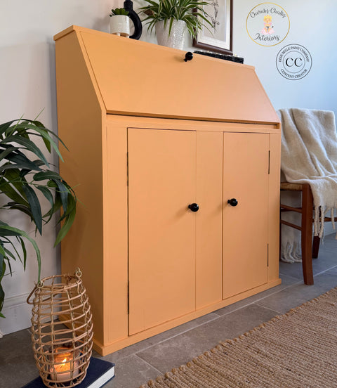 An orange-colored wooden write-on cabinet with a slanted top and two doors, styled in a home interior next to a chair, plants, and a woven basket candle holder.