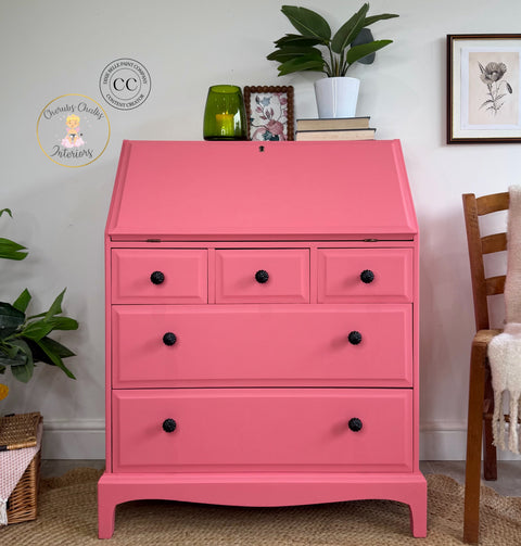 Bright pink painted secretary desk with black floral knobs, styled with books, greenery, framed artwork, and decorative accents in a cozy interior.