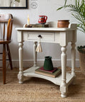 A vintage-style cream side table with a decorative tassel handle, holding a lit candle, red mug, and small brown container. Below, a green pitcher rests on stacked books. The setup is on a textured rug with a chair and potted plant nearby.