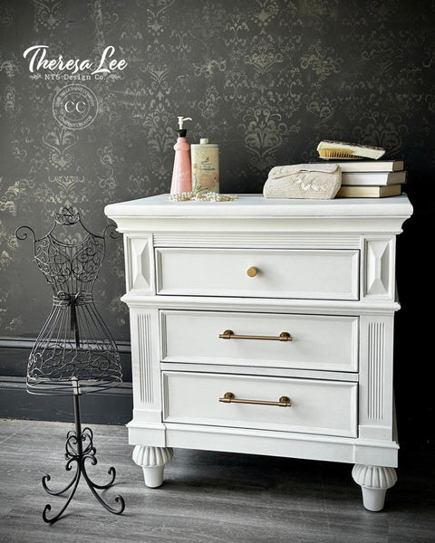A white vintage nightstand with gold handles stands against a dark, ornate wallpaper. On top are books, a purse, and toiletries. A decorative black wire mannequin stands to the left. "Theresa Lee NTS Design Co." watermark is in the corner.
