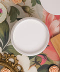 A round container filled with white paint sits on a floral-patterned surface, surrounded by decorative items including a gold frame, flowers, and a china saucer with a pink flower design.