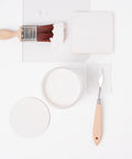 A paintbrush with white paint, a round container of white paint, a palette knife, and a square and circle of white paint samples are arranged neatly on a white background.