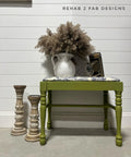 Green painted bench makeover with white and blue upholstery on the seat. Staged with an old camera and a vase of cattails on top. Two large candlesticks to the left of the bench.