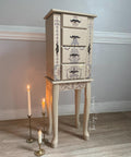 A vintage-style beige jewelry cabinet with intricate floral and cherub designs stands on long legs. Three lit white candles in brass holders are placed on the floor beside it. The cabinet is positioned against a light-colored wall.