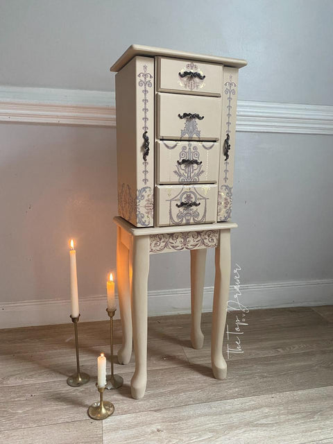 A vintage-style beige jewelry cabinet with intricate floral and cherub designs stands on long legs. Three lit white candles in brass holders are placed on the floor beside it. The cabinet is positioned against a light-colored wall.