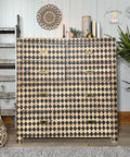 A dresser with a black and beige harlequin pattern stands against a white paneled wall. It is flanked by a decorative ladder with lights and a tall plant. A candle, books, and decor items are on top. Wall art and a logo are visible above.