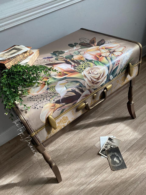 A vintage suitcase repurposed as a bench, adorned with floral artwork on top. A small plant and stacked books sit on the bench, with old photographs scattered on the wooden floor nearby.