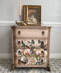An ornate dresser with floral designs sits against a light gray wall. It has four drawers, decorative handles, and is topped with a framed portrait, candles, and a small bird statue. Scattered papers lie on a patterned floor.