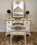 A white and gold vintage vanity with a matching chair sits on a wood floor. The vanity has ornate details, a large mirror, and two small topiary plants. A tufted bed is reflected in the mirror. Text reads, â€œMad for Rehab by Aleshia.â€.